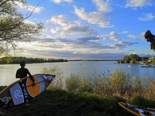 Ein junger Mann mit SUP-Board am Ufer des Alperstedter Sees. Der Himmel ist blau mit weißen Wolken, die Sonne scheint.