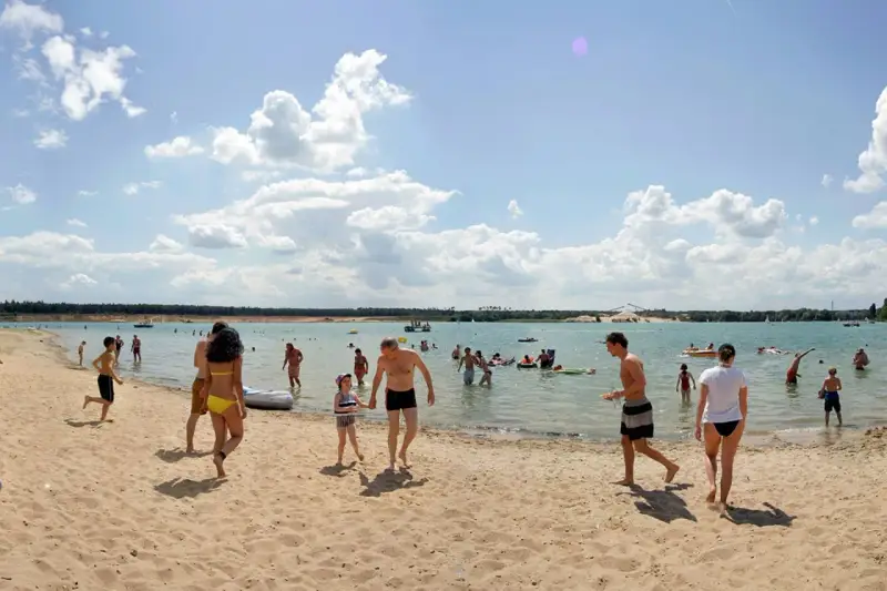 Menschen genießen einen sonnigen Tag am Badesee Frankfurt. Kinder planschen im Wasser, während Erwachsene sich am Sandstrand entspannen.